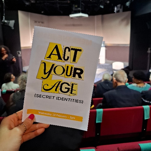 A programme with 'Act Your Age' on the front, held in front of a stage