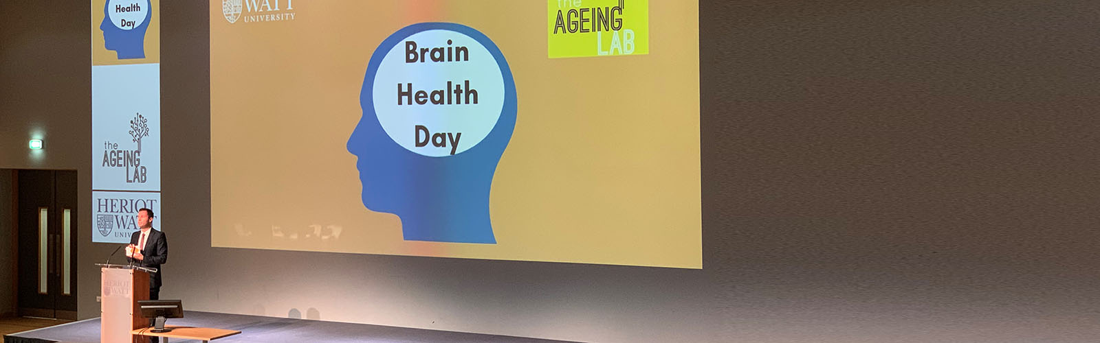 Professor Alan Gow from Heriot-Watt University stands in front of a screen with the headline 'Brain Health Day' showing